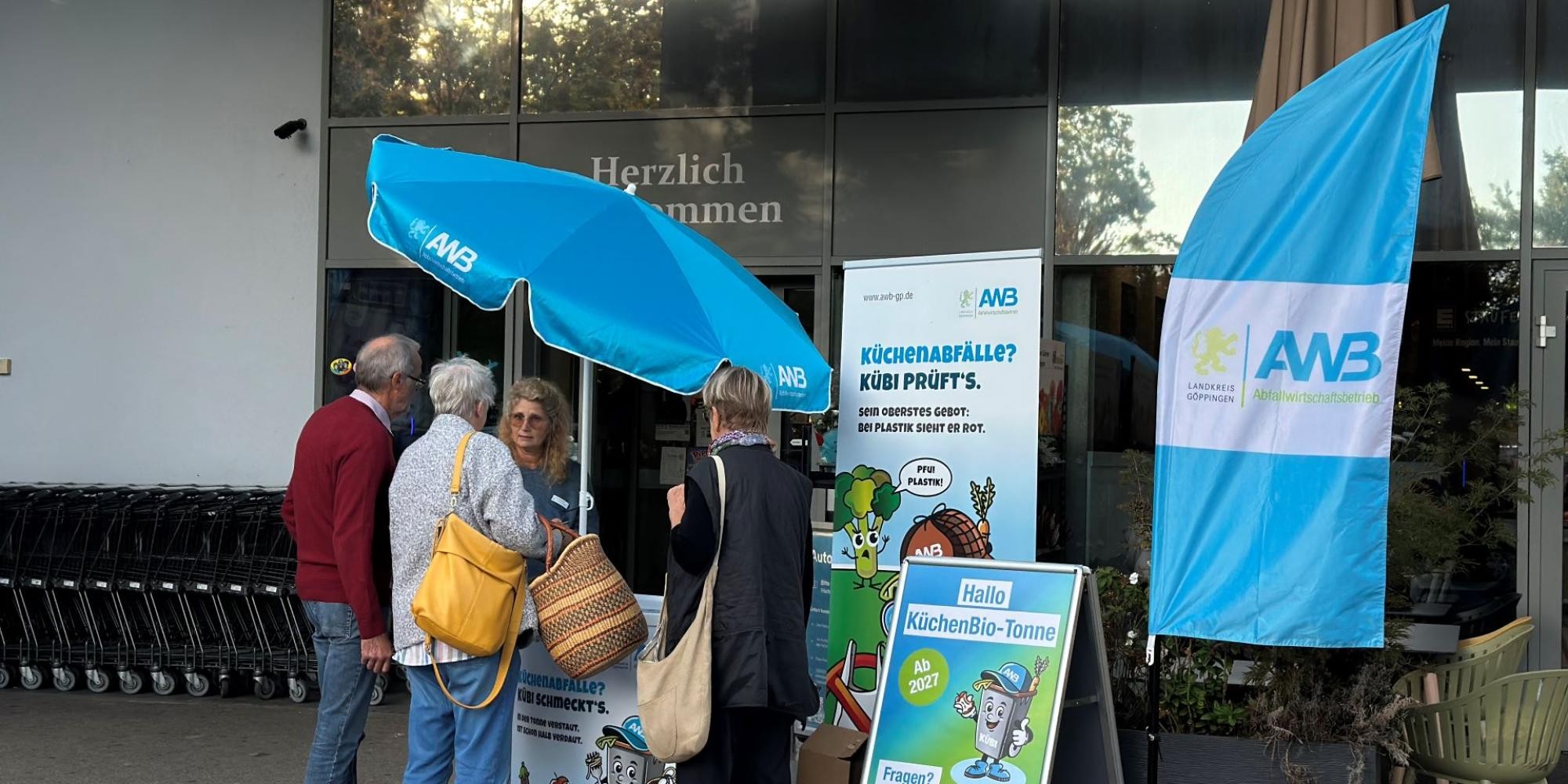 Vor dem Eingang eines Einkaufsmarktes steht ein Infostand mit AWB-Schirm, daneben AWB-Beachflag, Roll-up und Aufsteller zur KüchenBio-Tonne. Am Stand informiert eine Mitarbeiterin des AWB zwei Bürgerinnen und einen Bürger.