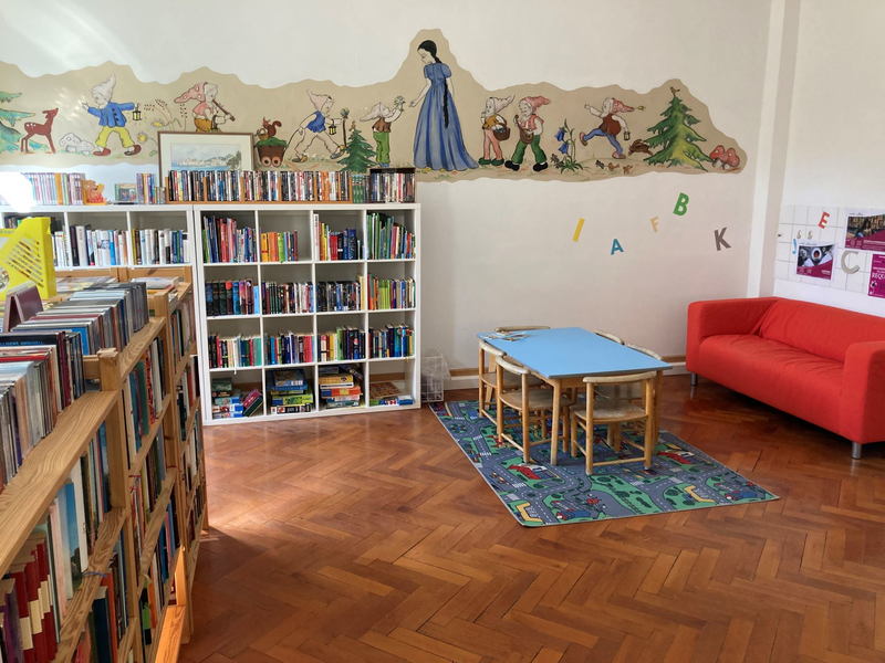 In einem gemütlich eingerichteten Raum stehen mehrere gefüllte Bücherregale. Rechts befindet sich ein rotes Sofa, davor ein Kindertisch mit Stühlen. 